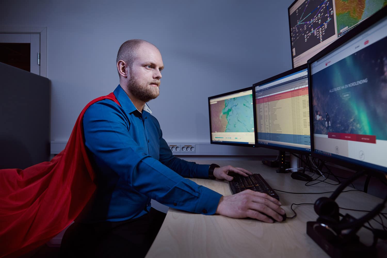 Børge fra IT hos Signal Bredbånd er ikledd rød kappe mens han jobber foran sine fem dataskjermer
