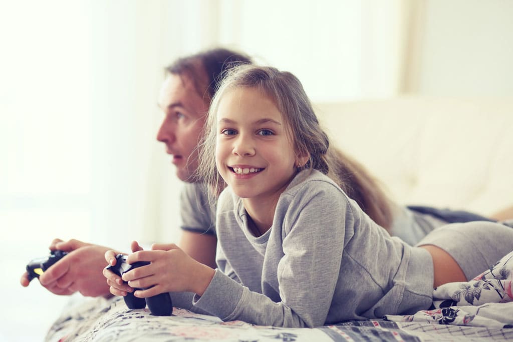 Jente ser inn i kameraet, glad og fornøyd, midt i en dataspillkamp med sin far, som er dypt konsentrert i bakgrunnen.