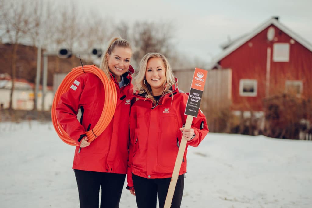 To damer fra Signal bredbånd i røde jakker holder fiberkabel og skilt foran hus om vinteren.