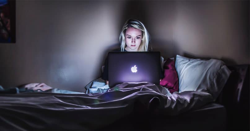 Blond jente sitter i senga i mørkt rom med en lysende laptop på fanget 