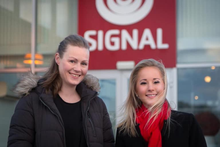 Økonomisjef Grethe Simonsen og leder for Markedsstøtte og Samfunnskontakt, Stine Værang står foran et rødt Signal-skilt.