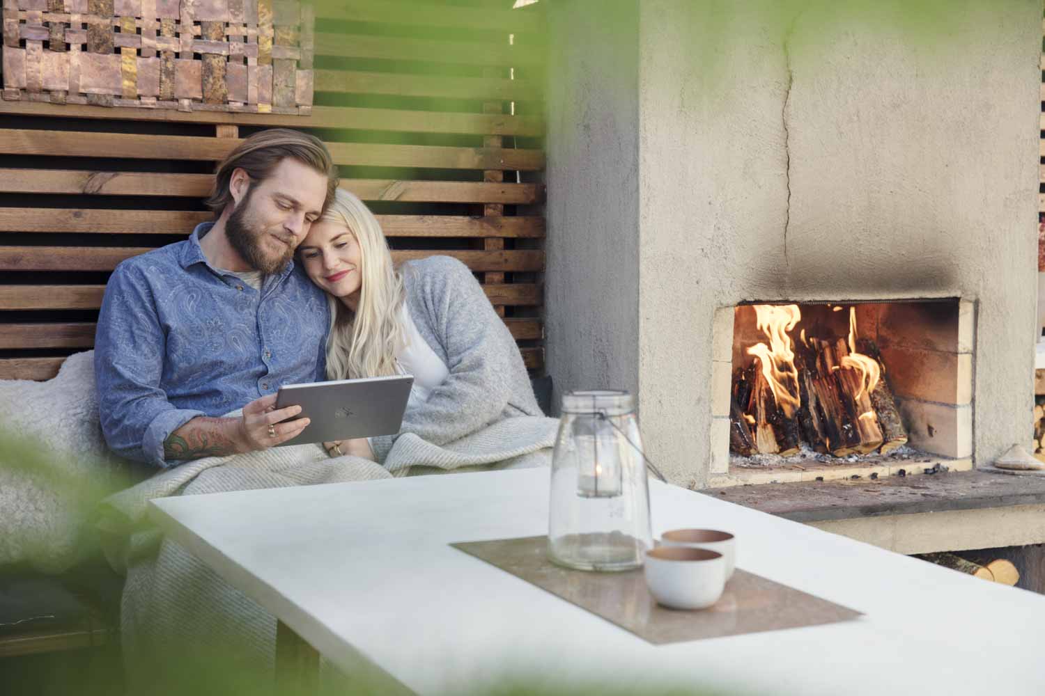 Jente sitter i armkroken til kjæresten sin og ser tv på ipad foran seg, ved siden av en utepeis.