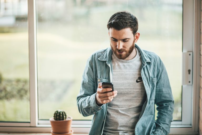 Mann i dongeriskjorte sitter i vinduskarmen mens han surfer på mobilen