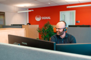 Kundesenterleder, Truls Are Ratkje sitter fremfor dataskjermer med headset på, og med rød vegg med signal logo i bakgrunnen