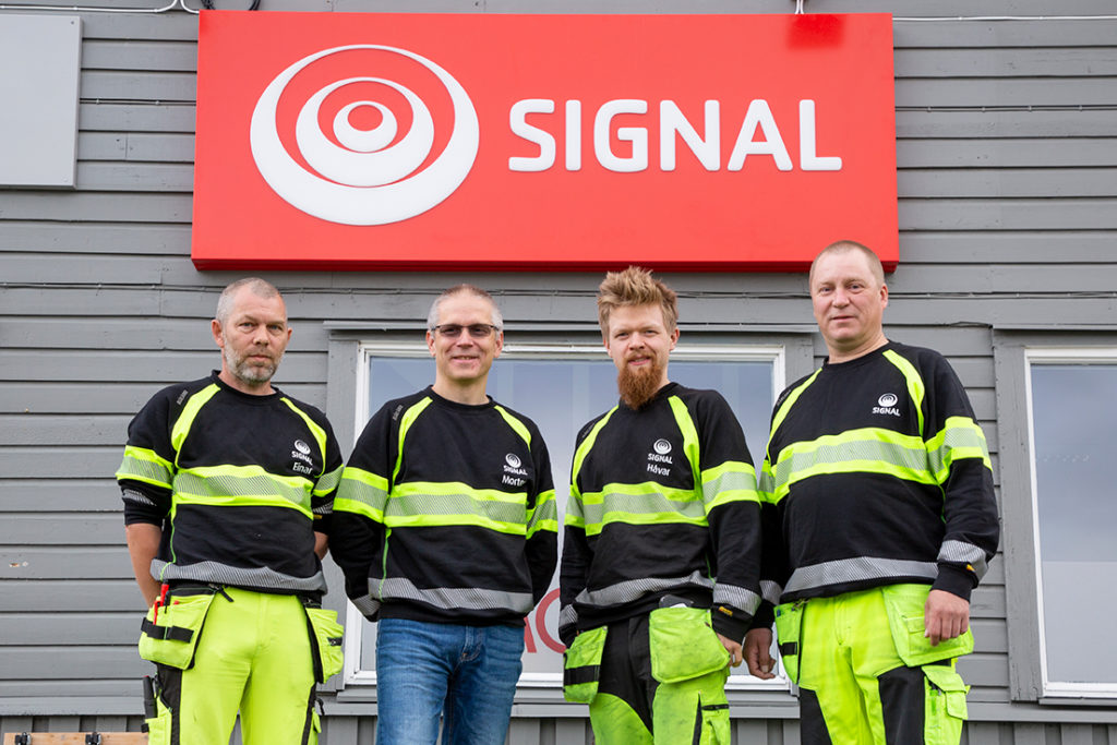 Fire menn kledd i svart og gult arbeidstøy står foran en grå vegg med rødt Signal-skilt