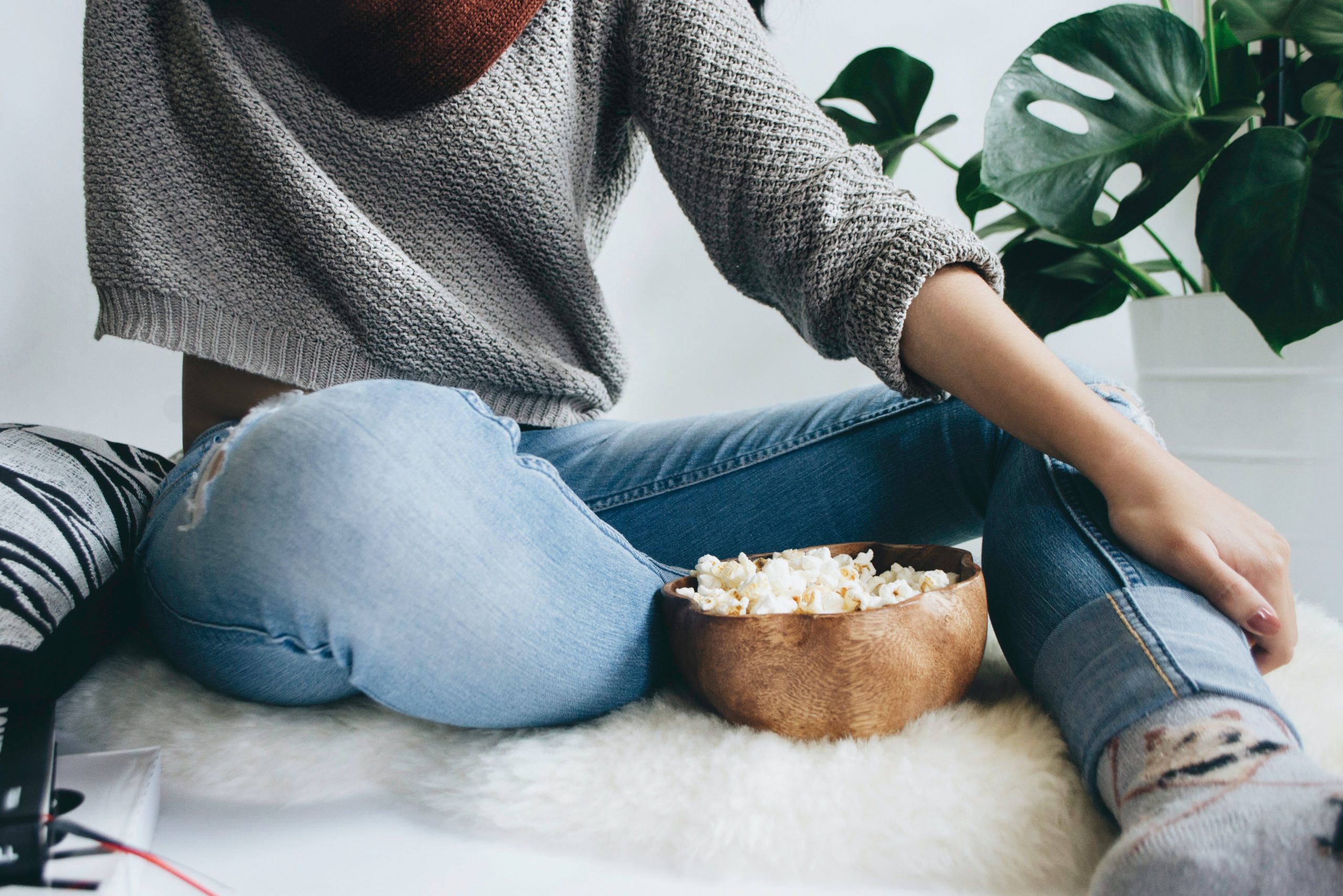 Dame i jeans og grå genser sitter på et hvitt skinn med en skål popcorn