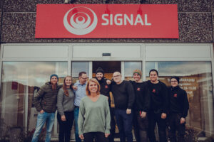 De ansatte i Signal Bredbånd avdeling Mosjøen står ved inngangspartiet under det røde Signal-skiltet.