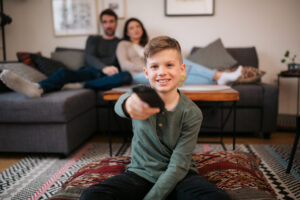 Smiling boy watching tv and changing the channel with a remote control with his parents on couch at the back. Caucasian kid enjoying watching television at home with parents relaxing on sofa behind him.